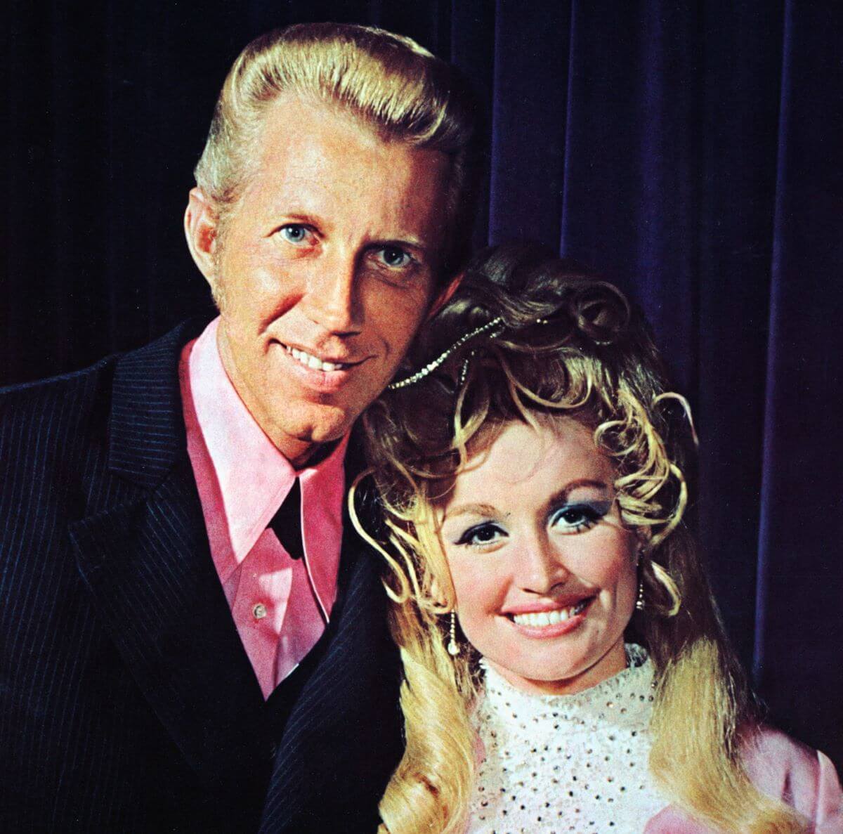Dolly Parton and Porter Wagoner smile together. They both wear pink shirts.