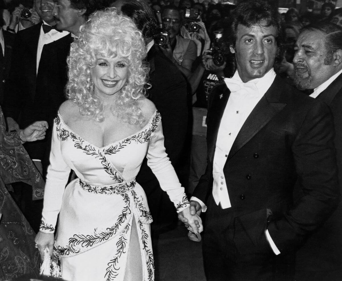 A black and white picture of Dolly Parton holding hands with Sylvester Stallone. She wears a dress and he wears a suit.