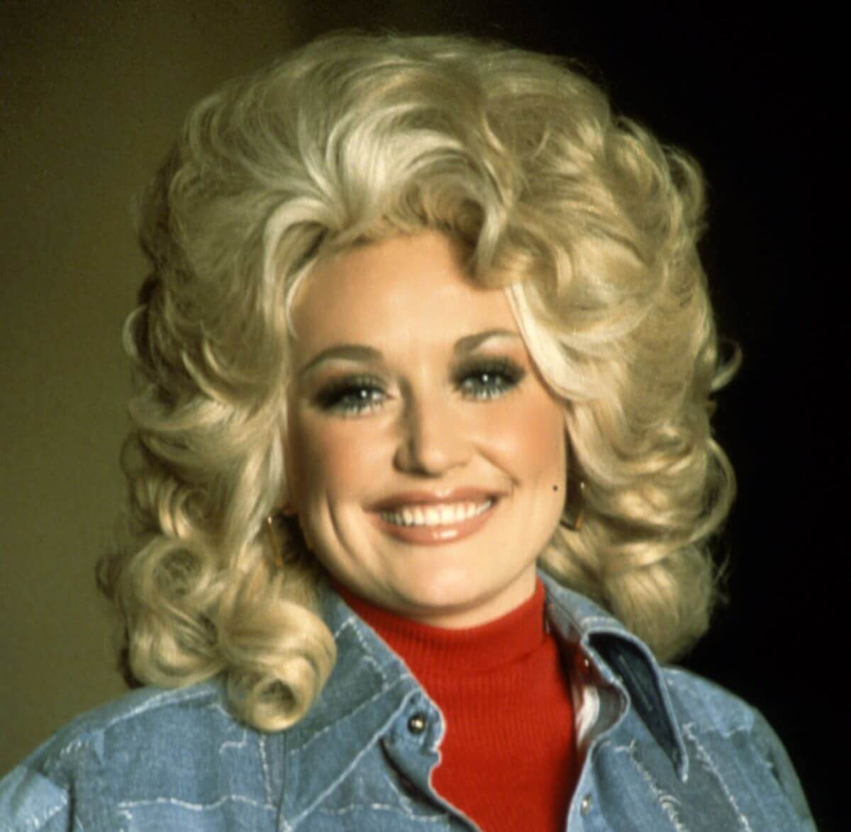 Dolly Parton wears a red shirt and a denim jacket. She smiles.
