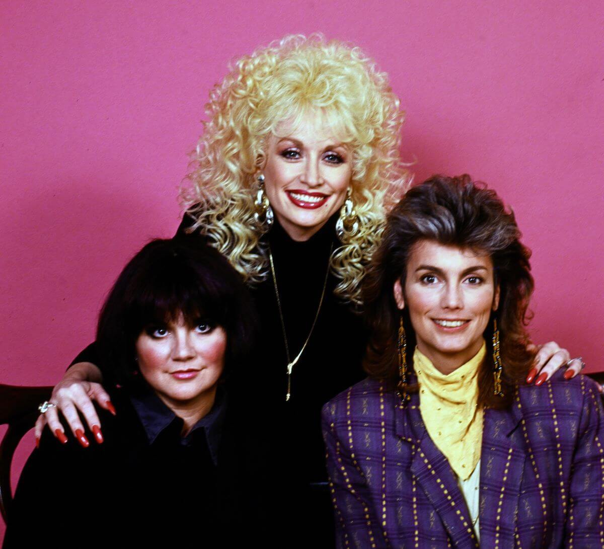 Dolly Parton stands behind Linda Ronstadt and Emmylou Harris. She has her hands on their shoulders.
