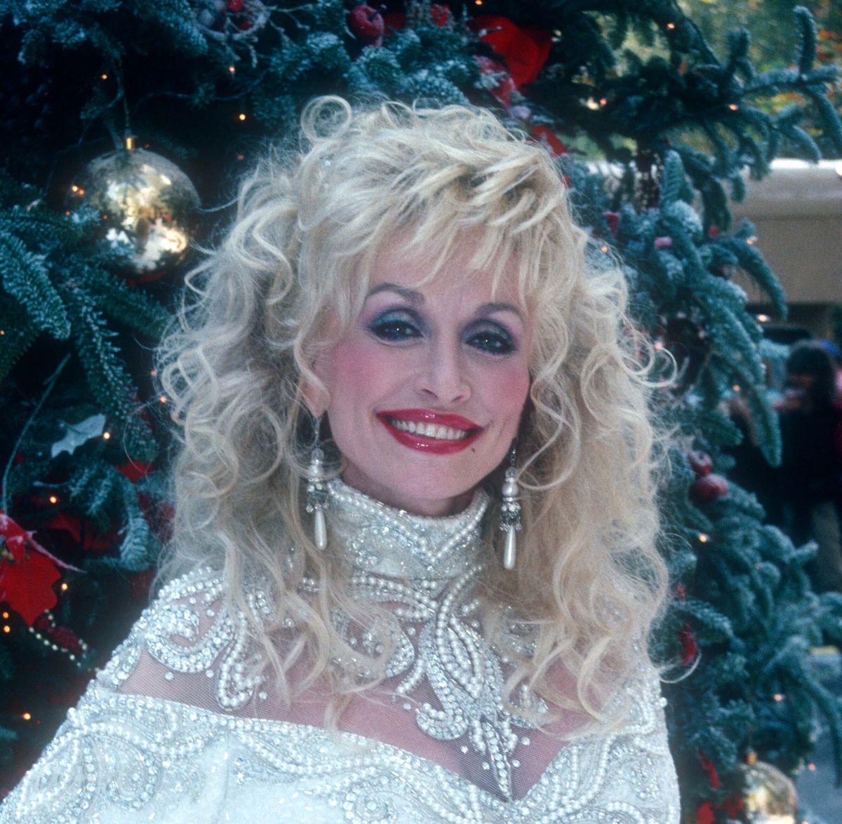 Dolly Parton wears a white dress and poses in front of a Christmas tree.