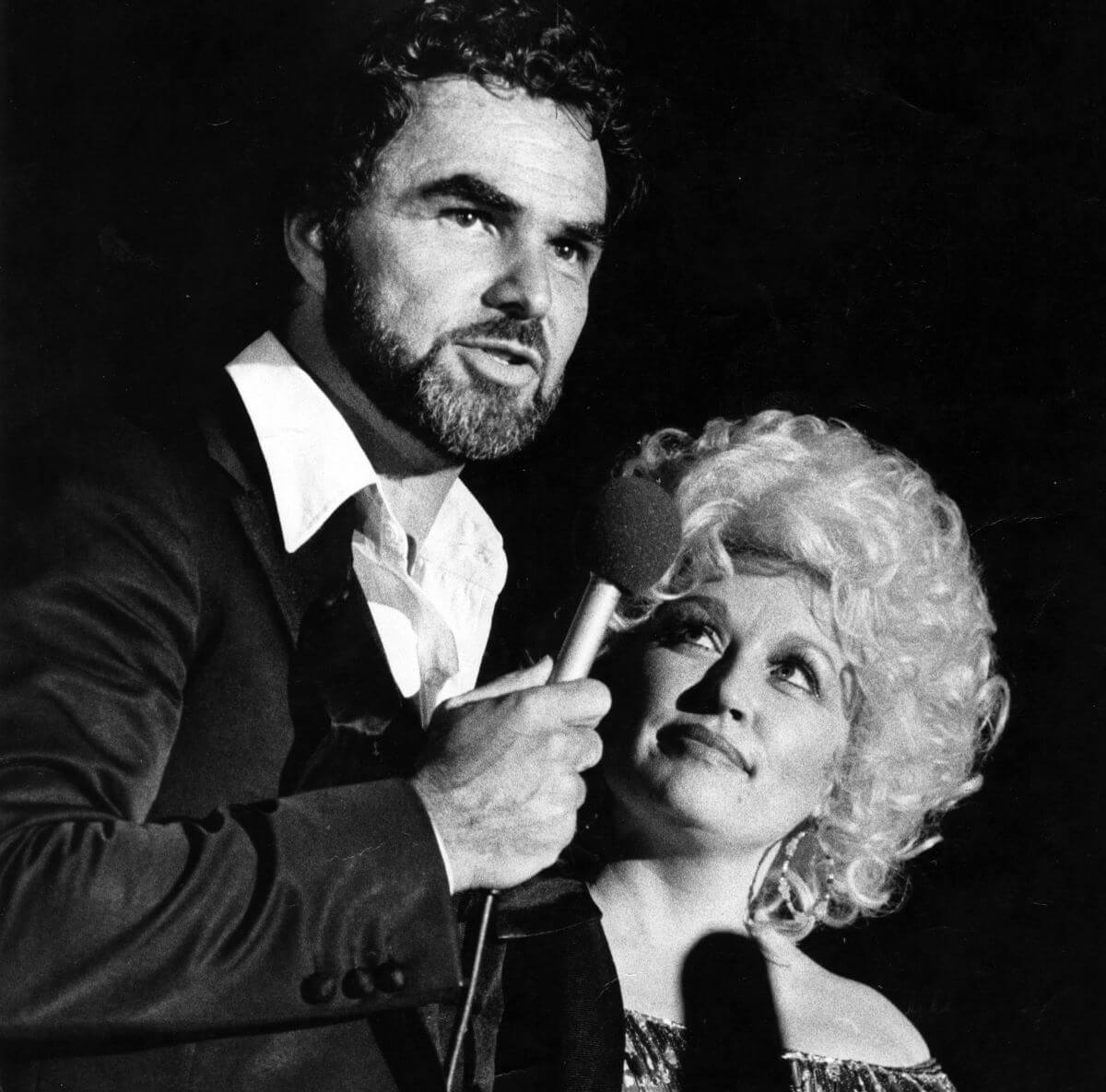 A black and white picture of Burt Reynolds holding a microphone while Dolly Parton looks up at him.
