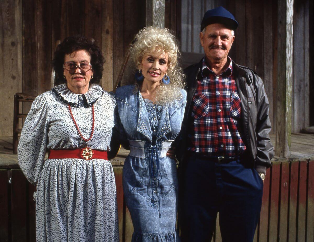 Dolly Parton wears a blue dress and stands between her mom and dad. They are in front of a cabin.