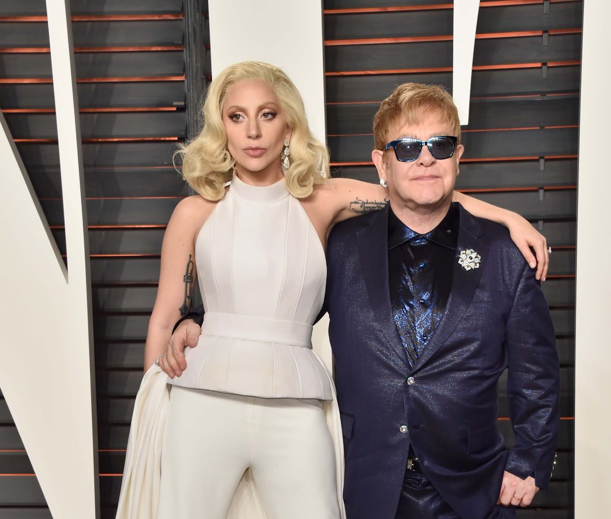 Lady Gaga wears white and stands with her arm around Elton John's shoulders. He wears a suit and sunglasses.