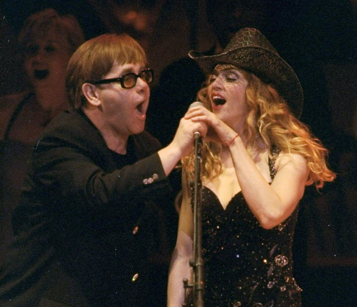 Elton John wears sunglasses and sings into a microphone with Madonna, who wears a black cowboy hat. They both hold one hand on the microphone.