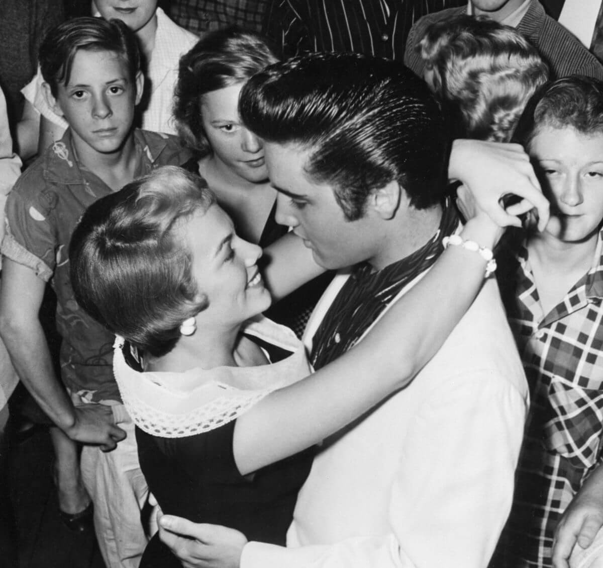 A black and white picture of Elvis dancing with his girlfriend, Anita Wood. They stand in a crowd of people.