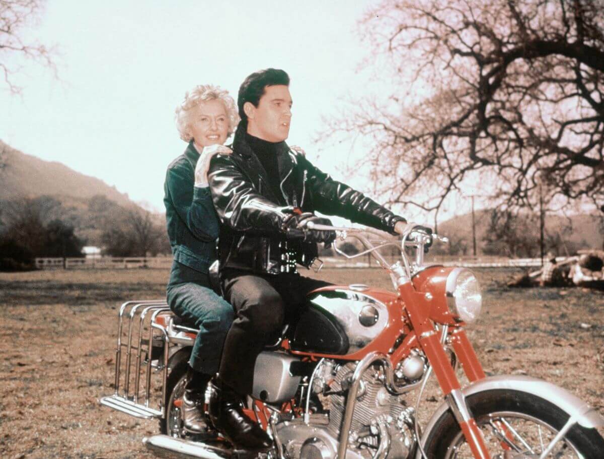 Elvis Presley rides a red motorcycle with Barbara Stanwyck behind him on the set of 'Roustabout.'