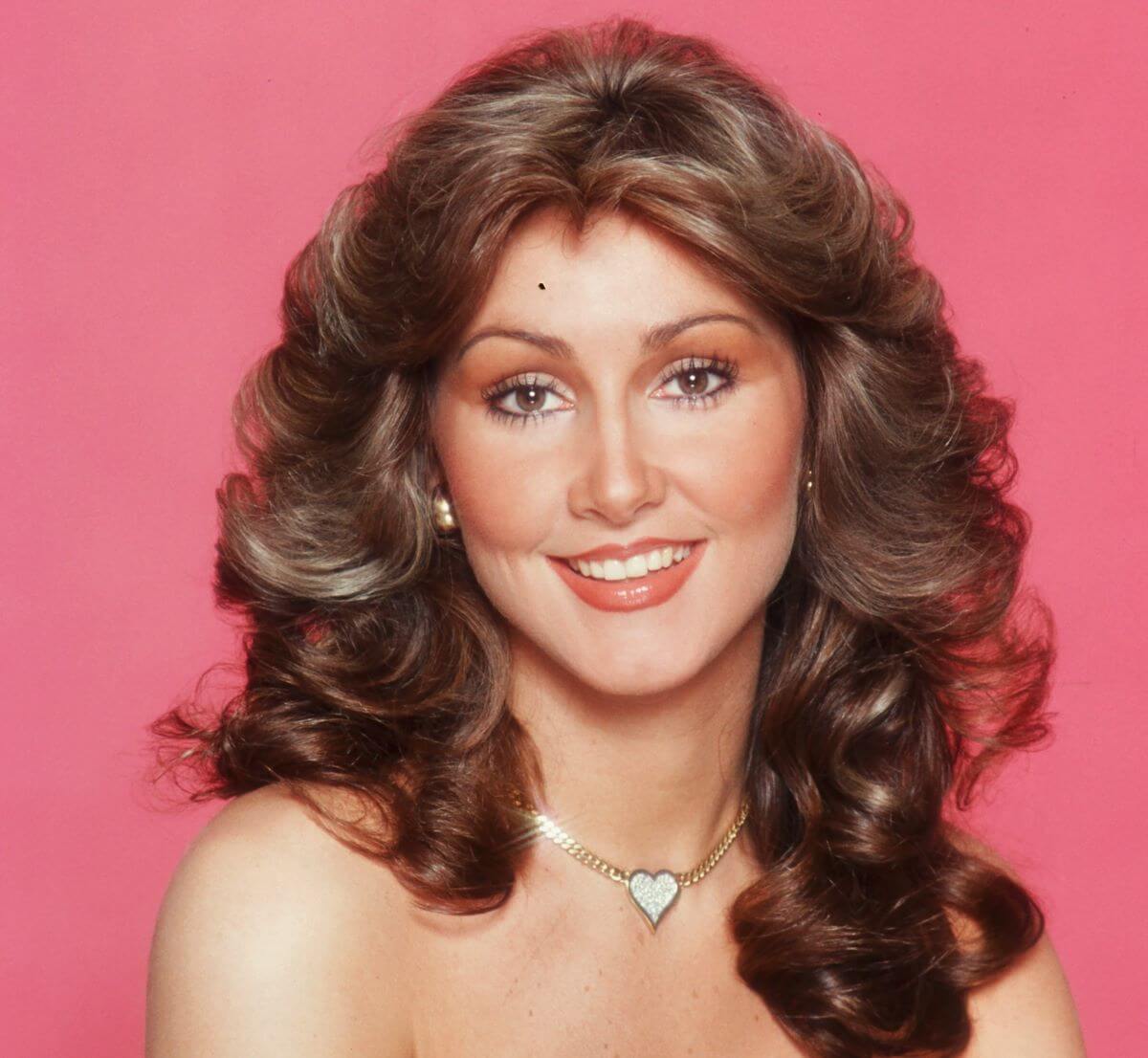 Linda Thompson smiles against a pink background. She wears a heart shaped necklace.