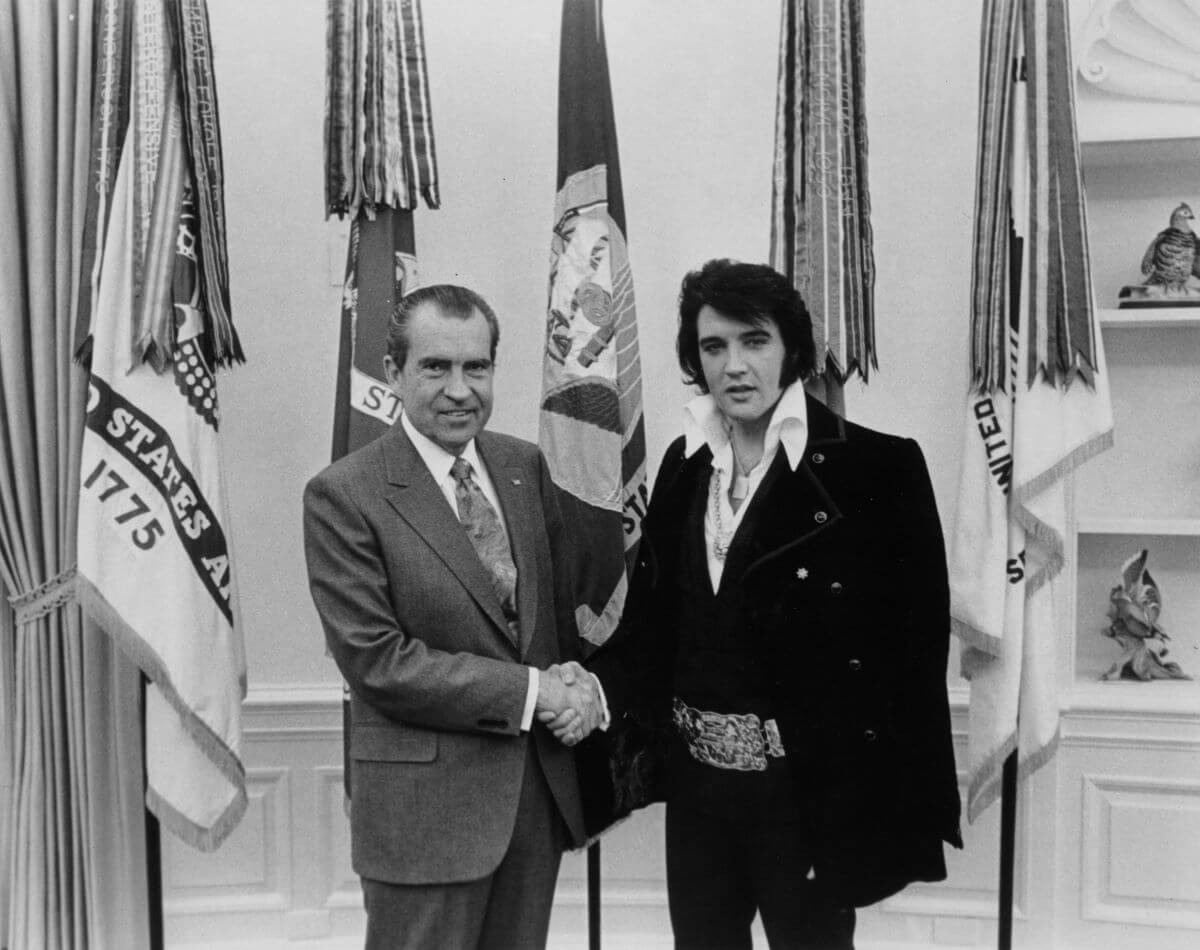 A black and white picture of Elvis and Richard Nixon standing in front of a row of flags and shaking hands.