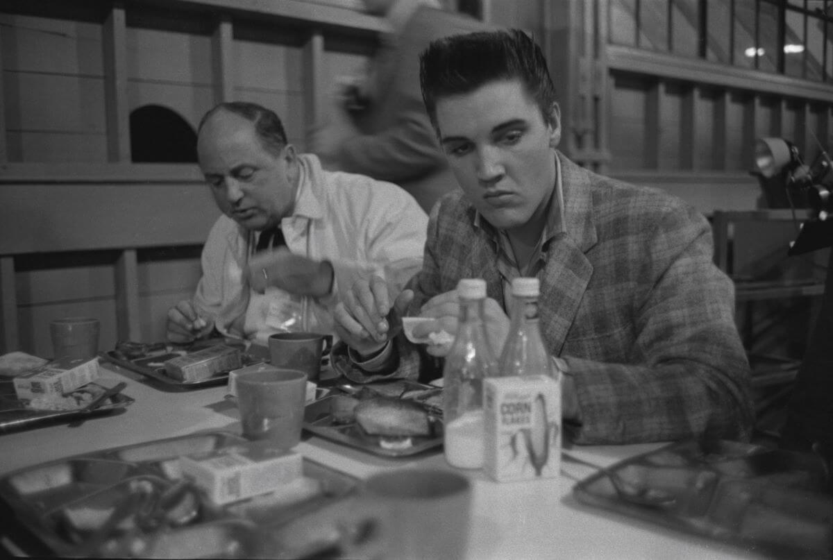 A black and white picture of Colonel Tom Parker and Elvis sitting on the same side of a booth and eating.