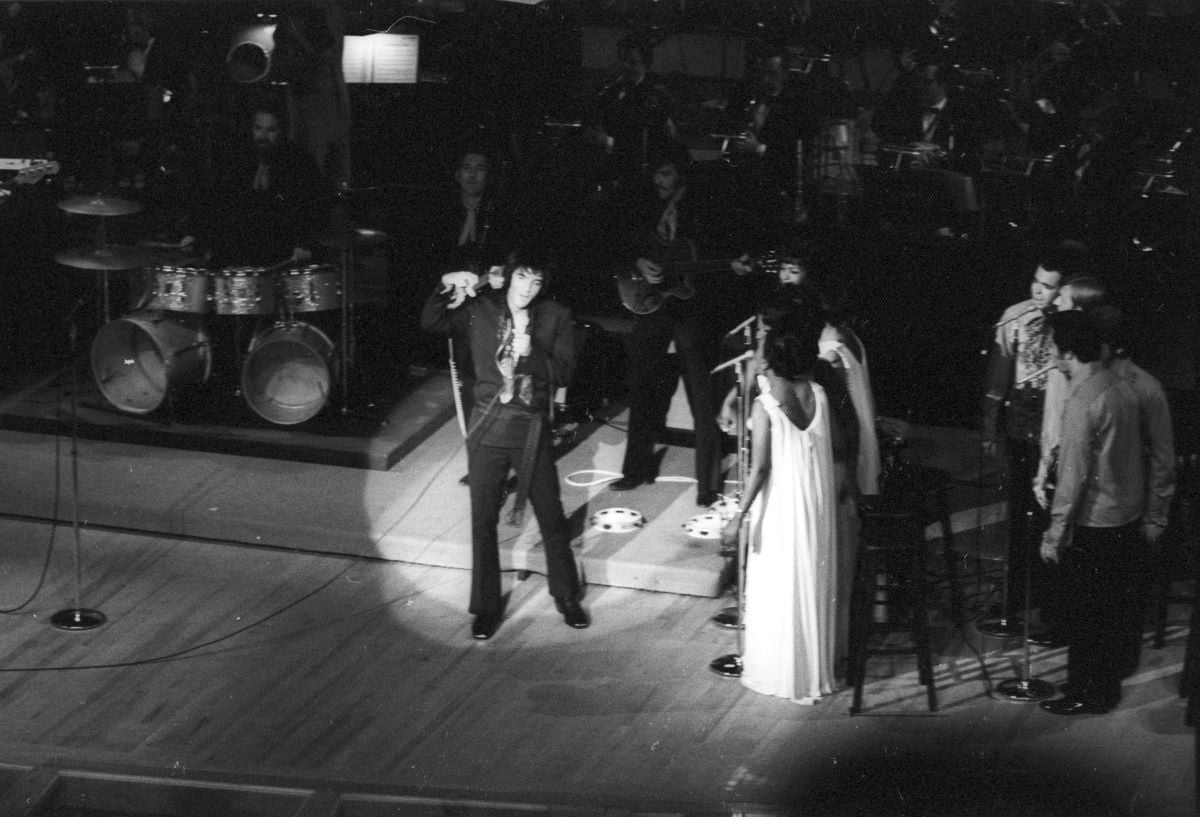 A black and white picture of Elvis onstage with the Sweet Inspirations. He stands in a spotlight.