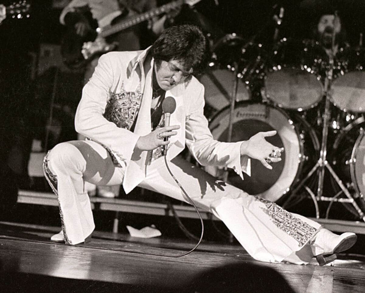 A black and white picture of Elvis in a deep lunge as he sings into a microphone.