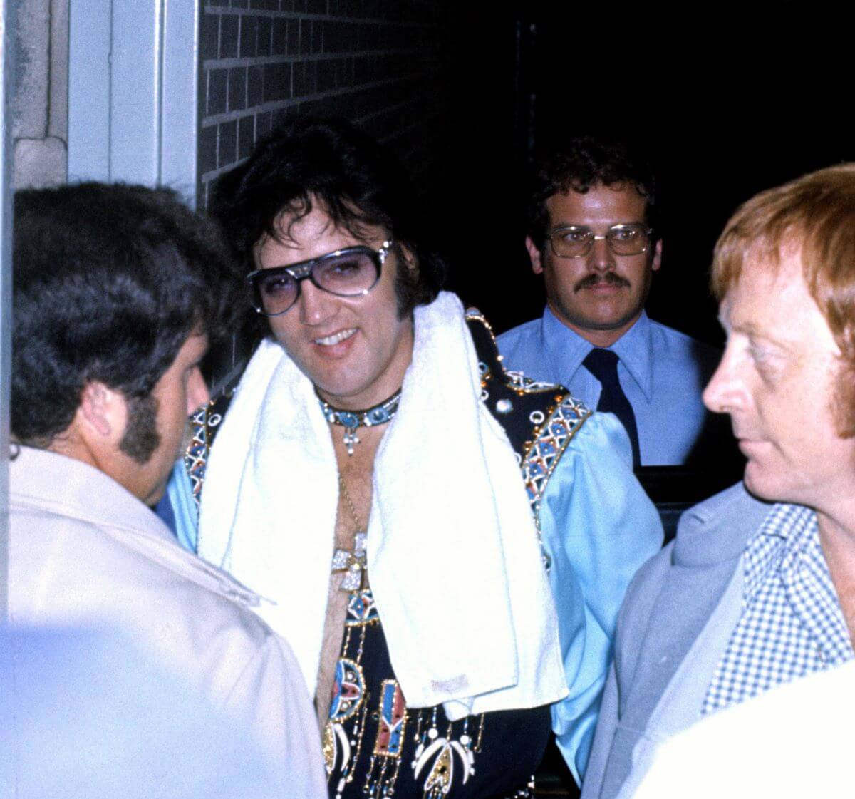 Elvis has a towel around his neck and wears sunglasses. He walks with his bodyguards.