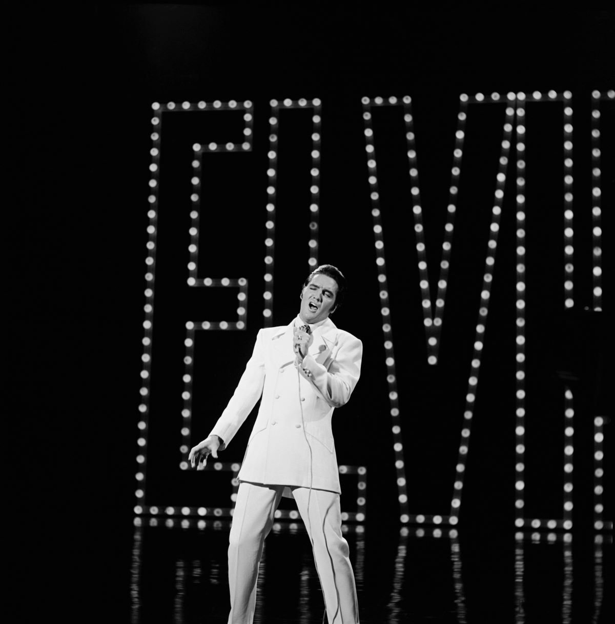 A black and white picture of Elvis Presley singing into a microphone in front of his name in lights.