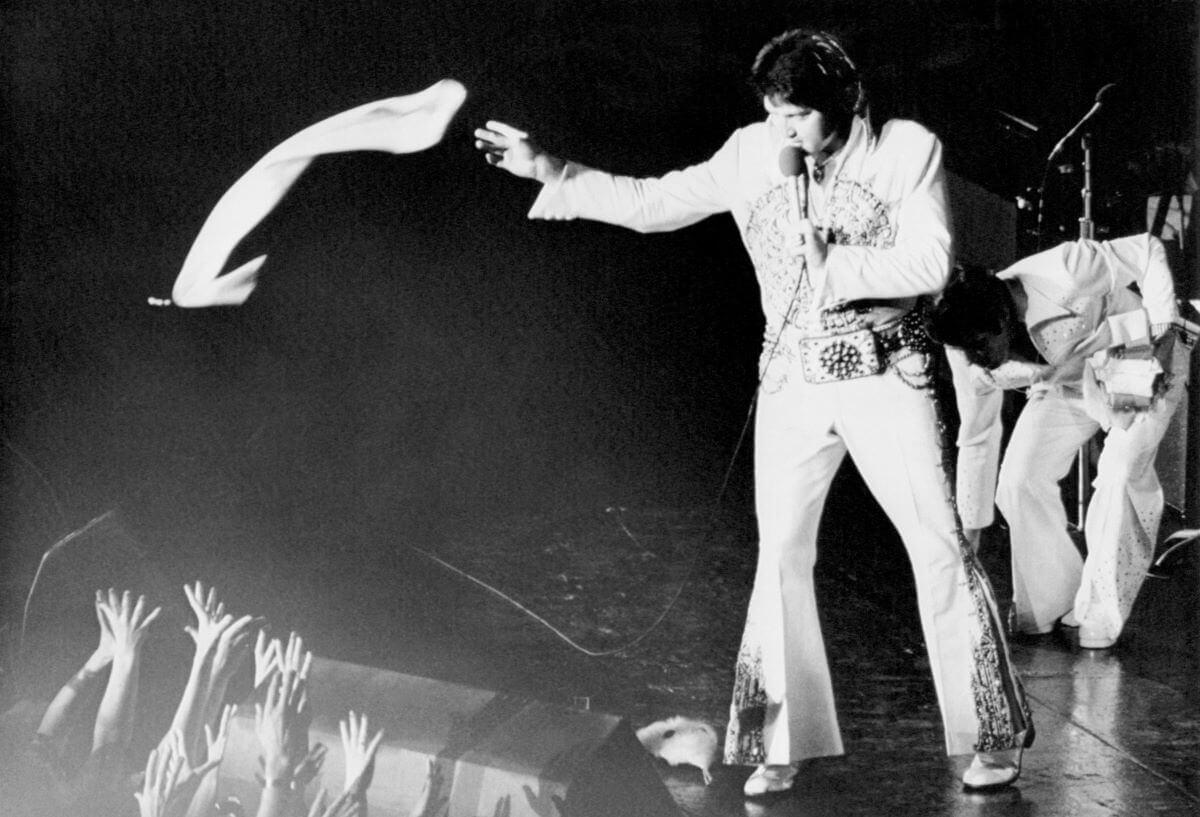 A black and white picture of Elvis Presley tossing a scarf to his audience during a concert. He holds a microphone.