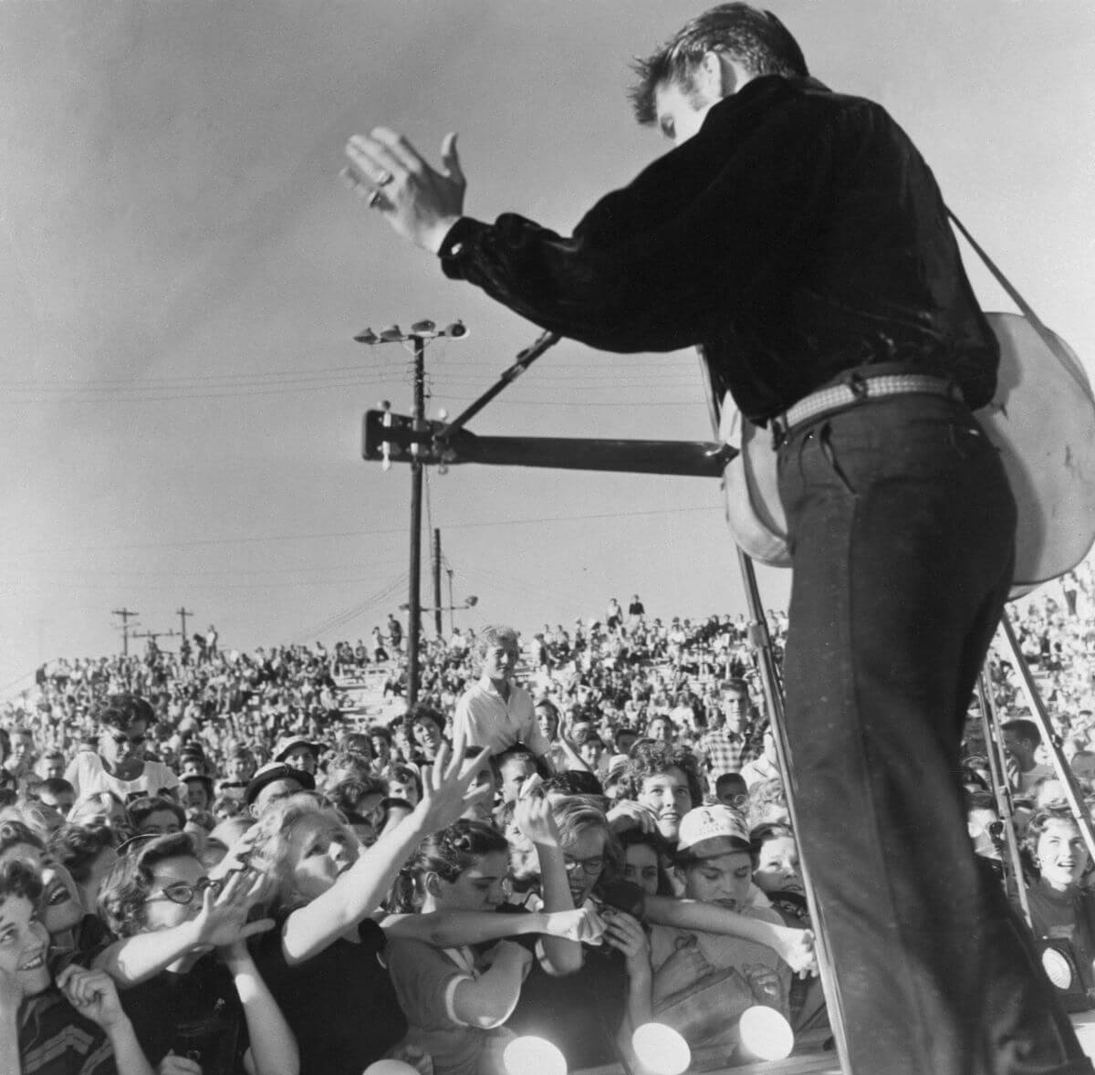 A black and white picture of Elvis Presley holding a guitar and waving at his audience.