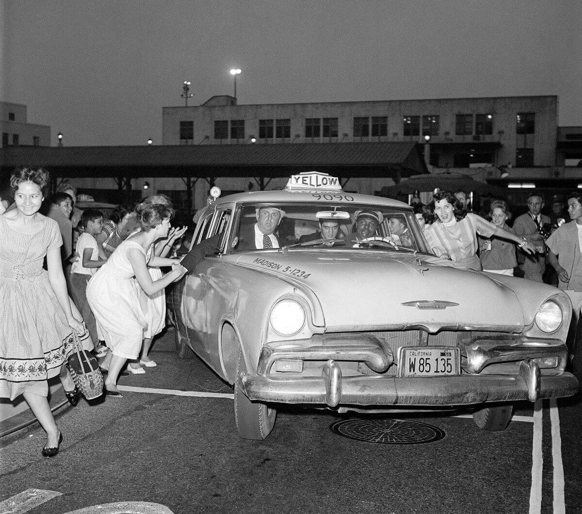 A black and white picture of Elvis fans chasing after him while he sits in a taxi.