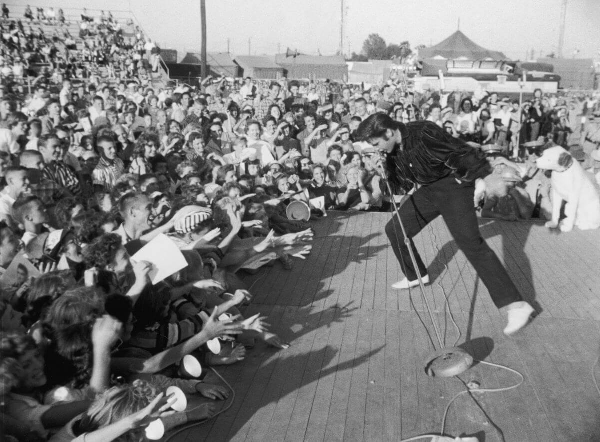Elvis sings into a microphone while fans reach towards her.