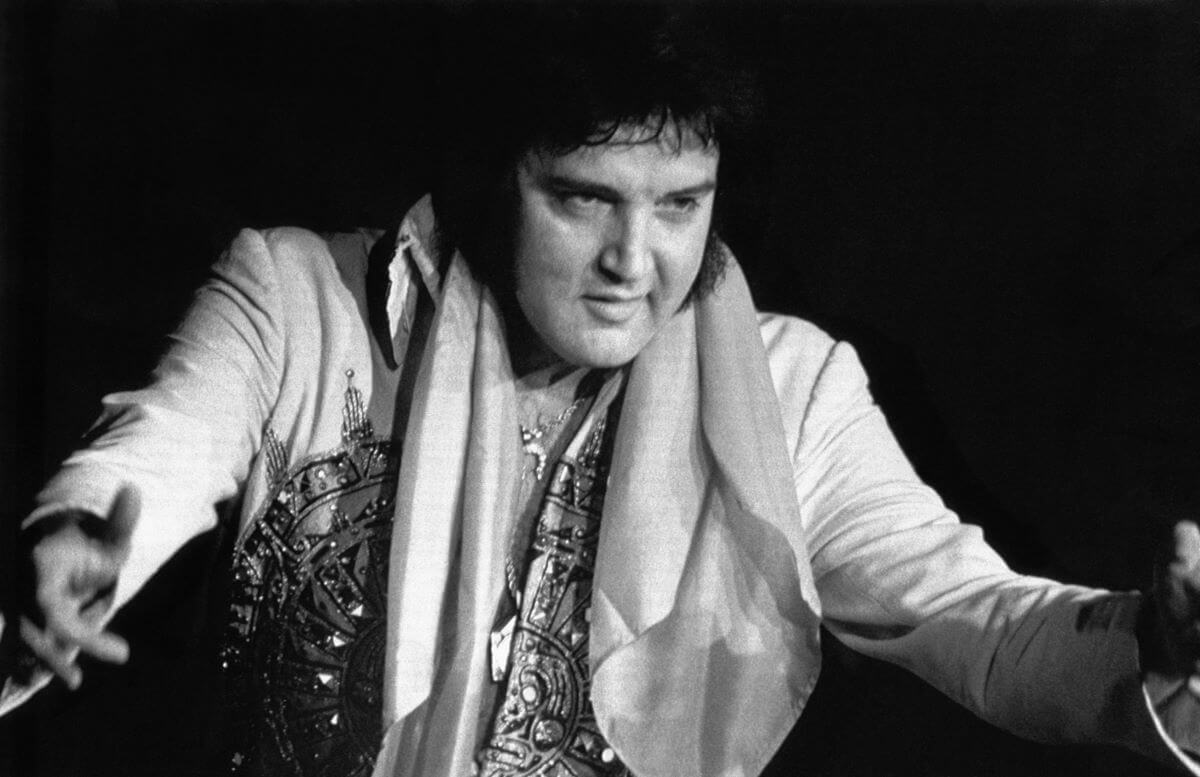 A black and white picture of Elvis wearing a jumpsuit and a scarf around his shoulders. He reaches his arms out while performing.