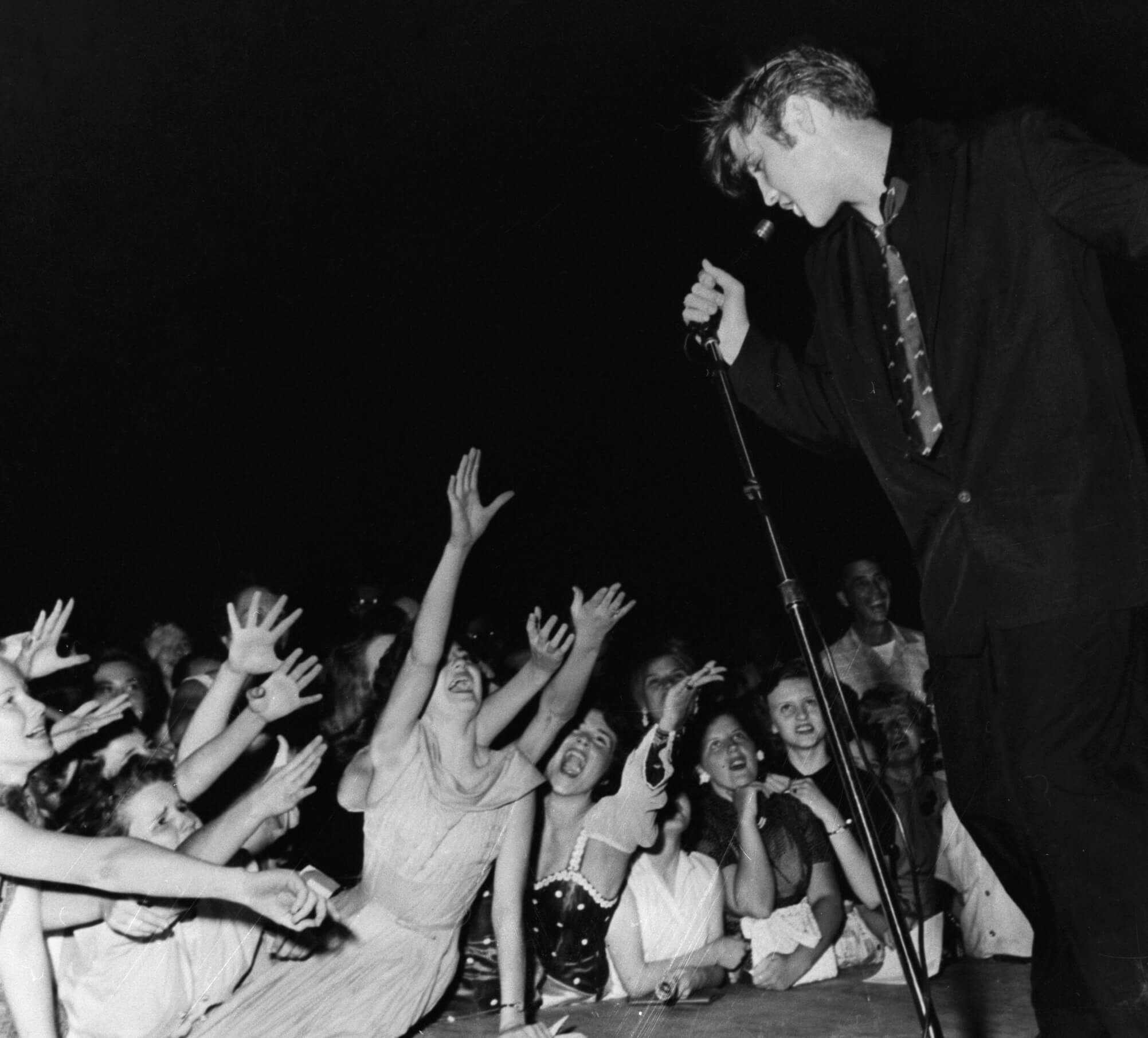 A black and white picture of Elvis standing onstage and singing to fans, who reach up towards him.