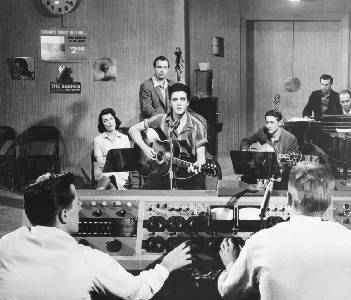 A black and white picture of Elvis playing guitar in the recording studio. He sits next to other musicians and in front of two audio technicians.