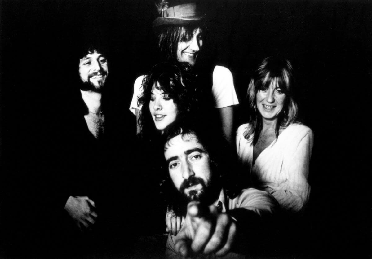 A black and white picture of Fleetwood Mac posed together. Mick Fleetwood wears a hat and stands in the back, Lindsey Buckingham, Stevie Nicks, and Christine McVie stand in the middle. John McVie stands in front and points at the camera.