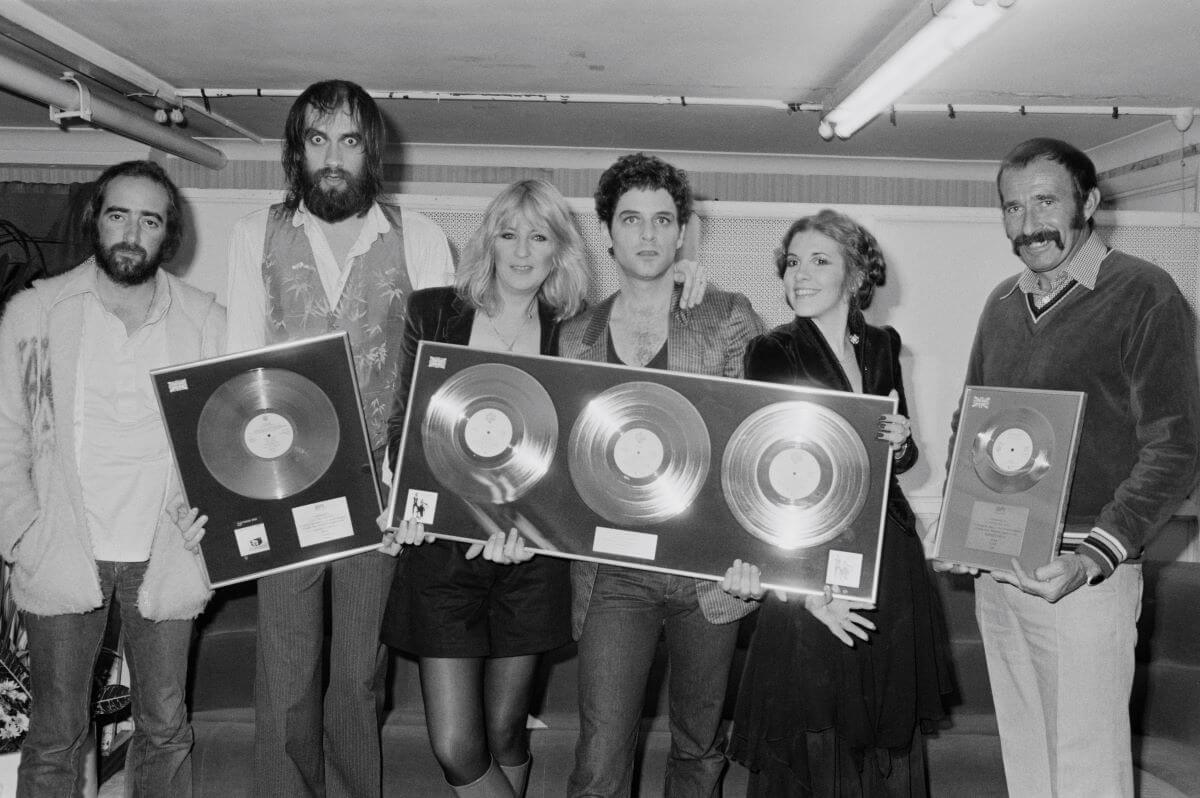 A black and white icture of John McVie, Mick Fleetwood, Christine McVie, Lindsey Buckingham, Stevie Nicks of Fleetwood Mac, and a man posing with framed records.