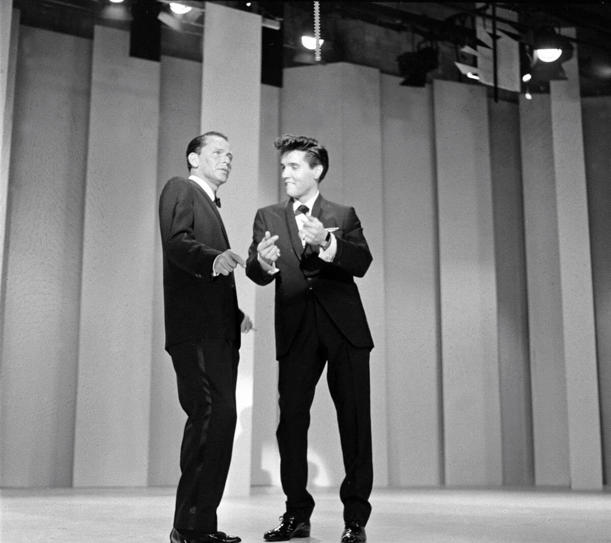 A black and white picture of Frank Sinatra and Elvis wearing tuxedos and dancing on a sound stage.