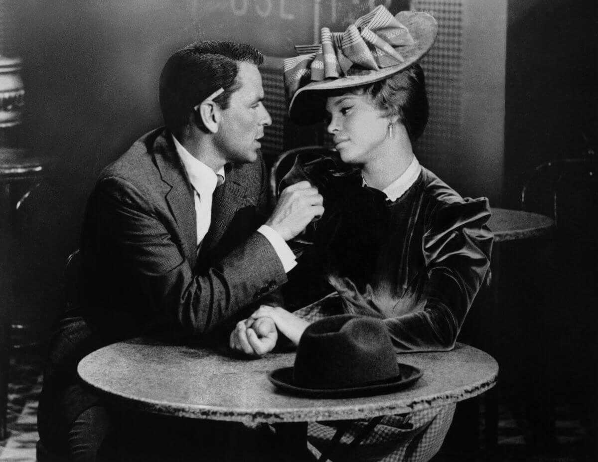 A black and white picture of Frank Sinatra and Juliet Prowse sitting at a table together in 'Can Can.' She wears a hat.