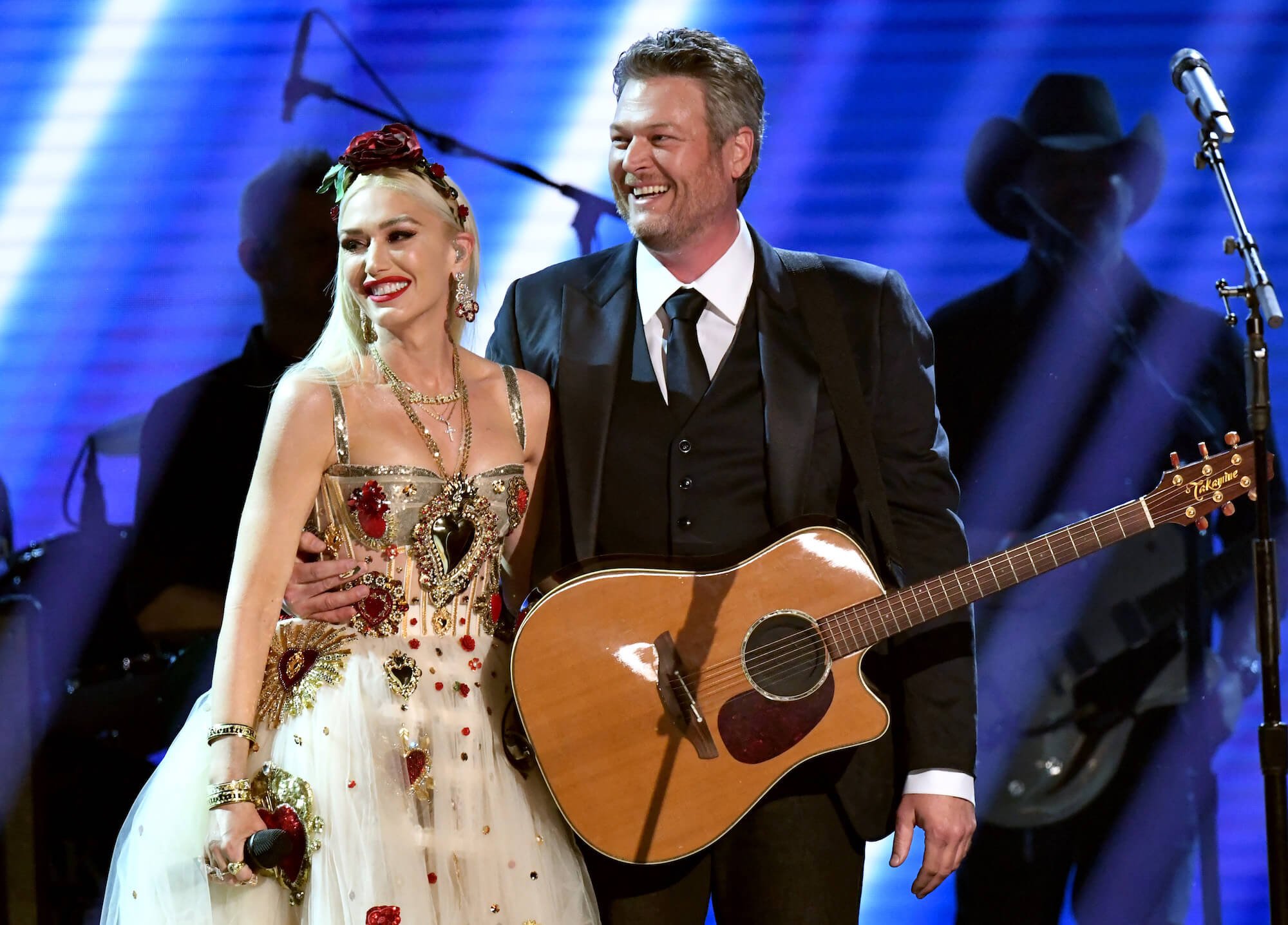 Gwen Stefani wearing a dress while standing next to Blake Shelton in a suit. They're smiling on stage. Shelton has a guitar strapped around his body.