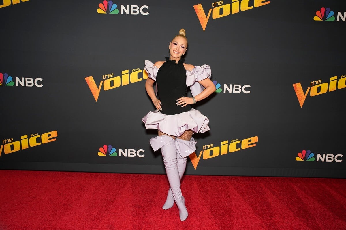 Gwen Stefani posing in front of a poster of 'The Voice' in a black and white dress.