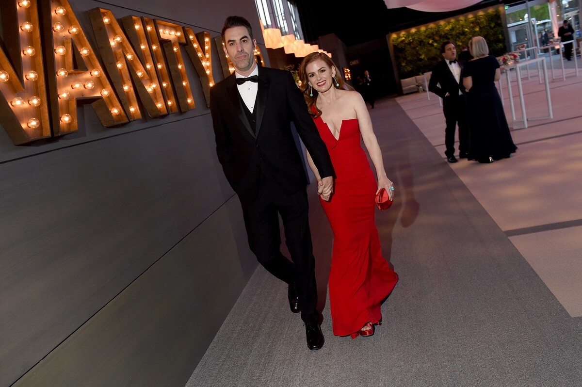 Sacha Baron Cohen wearing a suit while holding hands with Isla Fisher at the 2018 Vanity Fair Oscar Party.