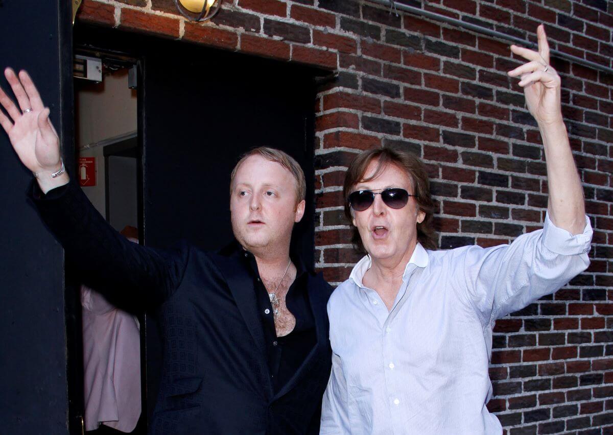 James McCartney and Paul McCartney stand next to each other and lift one arm in the air. Paul McCartney wears sunglasses.