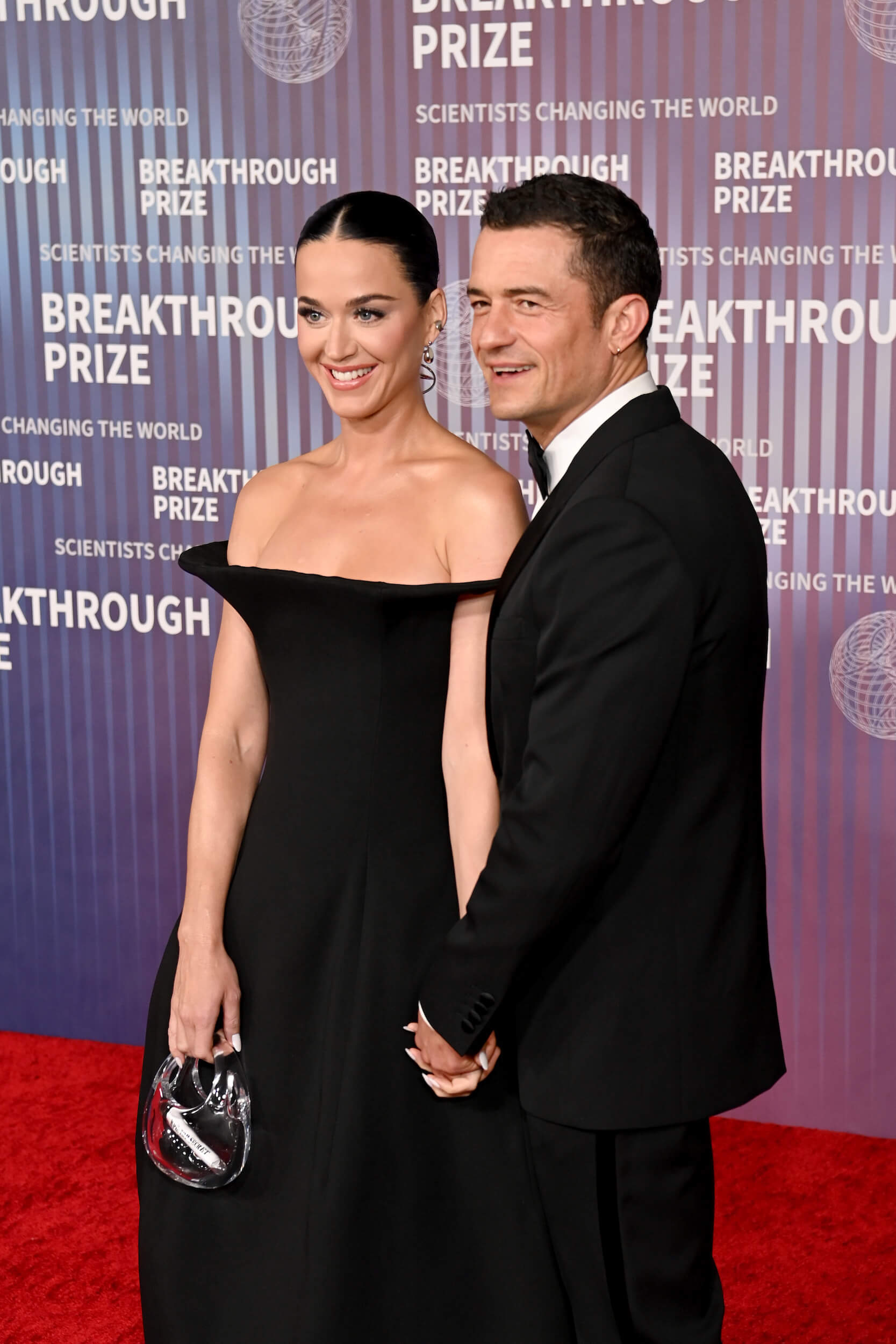 Katy Perry and Orlando Bloom standing on the red carpet while attending the 10th Annual Breakthrough Prize Awards. Perry is dressed in a black dress and Bloom is dressed in a tuxedo.
