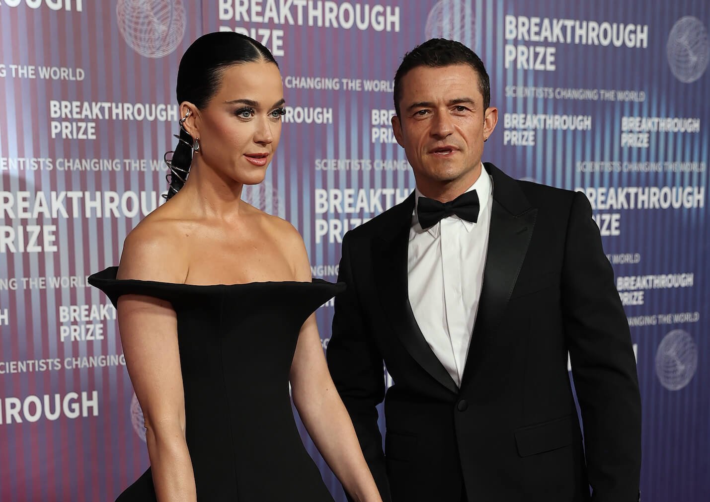 Katy Perry and Orlando Bloom holding hands at the Breakthrough Prize Awards. Perry is wearing a black dress and Bloom is wearing a tuxedo.