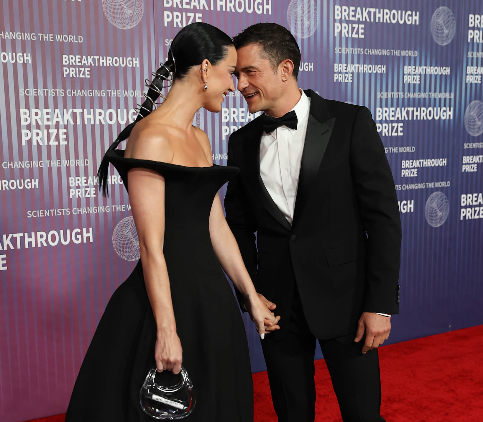 Katy Perry and Orlando Bloom standing on the red carpet while attending the 10th Annual Breakthrough Prize Awards. Perry is dressed in a black dress and Bloom is dressed in a tuxedo.