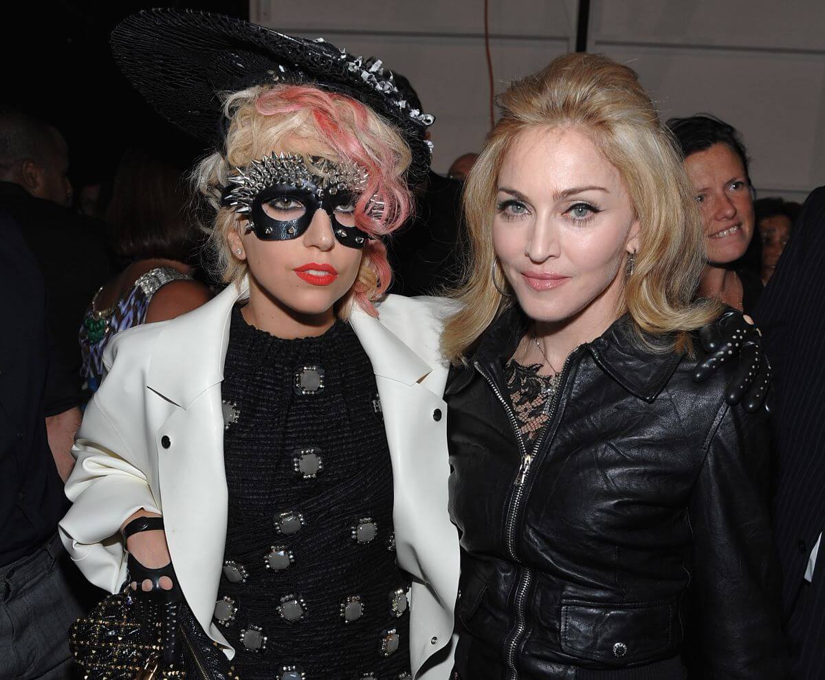 Lady Gaga wears a studded eye mask and hat. She stands with Madonna, who wears black.