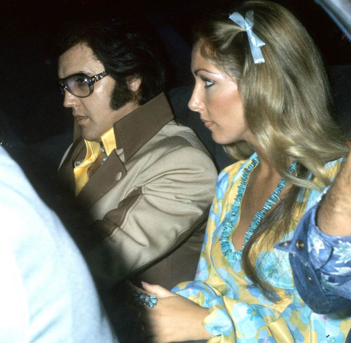 Elvis sits next to Linda Thompson. He wears sunglasses and she wears a blue necklace and blue ribbon in her hair.