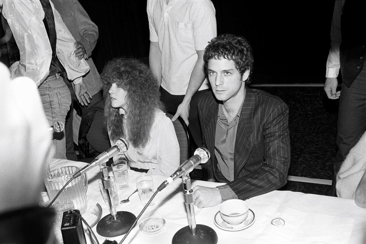 A black and white picture of Stevie Nicks and Lindsey Buckingham sitting in front of microphones on the same side of a table.