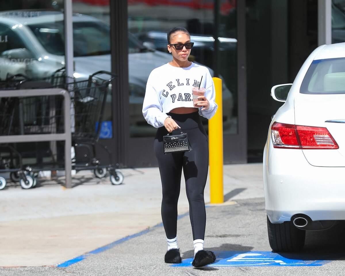 Model Lori Harvey carries a smoothie after working out in black leggings and a gray cut-off sweatshirt