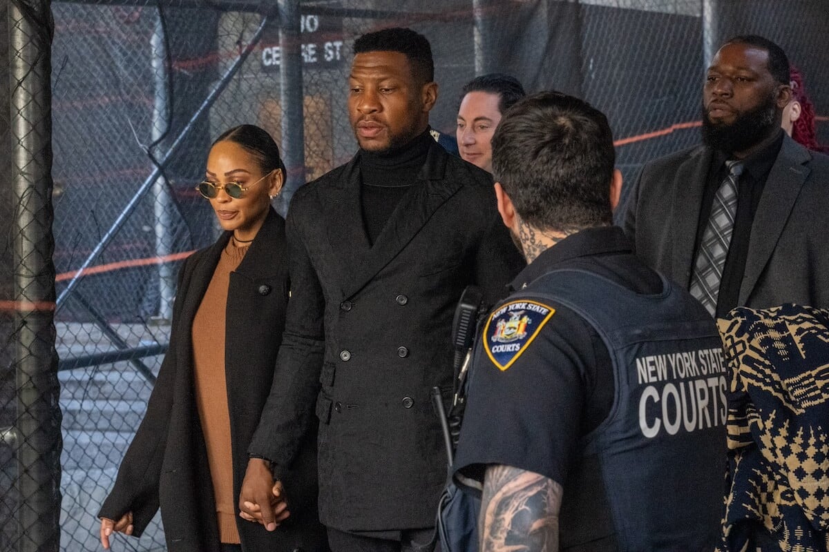 Jonathan Majors, in a black suit, holds hands with Meagan Good as they leave court