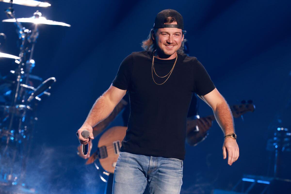 Morgan Wallen wears a black shirt and backwards black baseball hat. He holds a microphone.