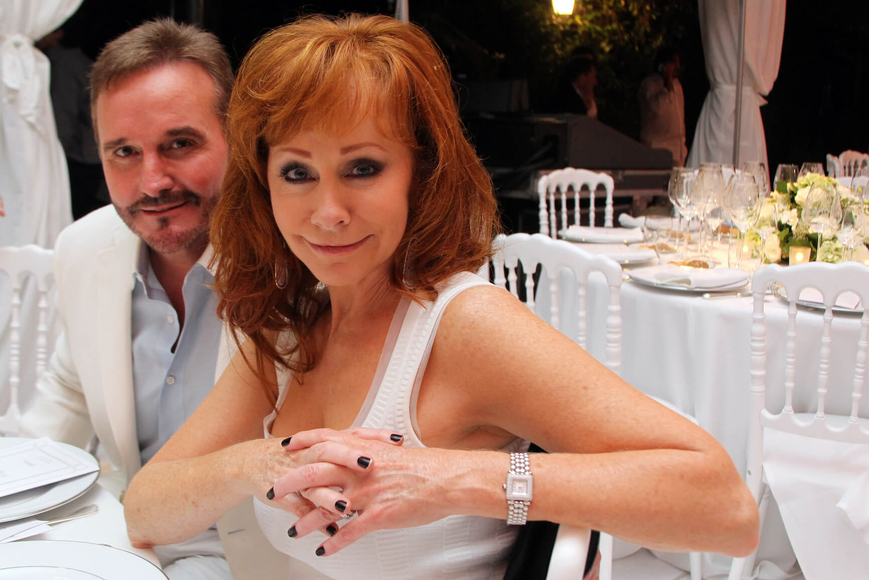 Narvel Blackstock sitting behind Reba McEntire at a white table in 2014. They are attending the White Party Dinner Hosted by Andrea and Veronica Bocelli.