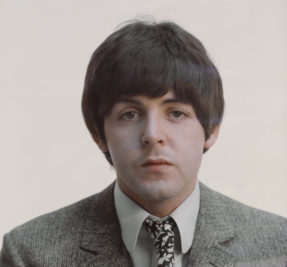 The Beatles' Paul McCartney wears a suit and tie and poses in front of a beige background.