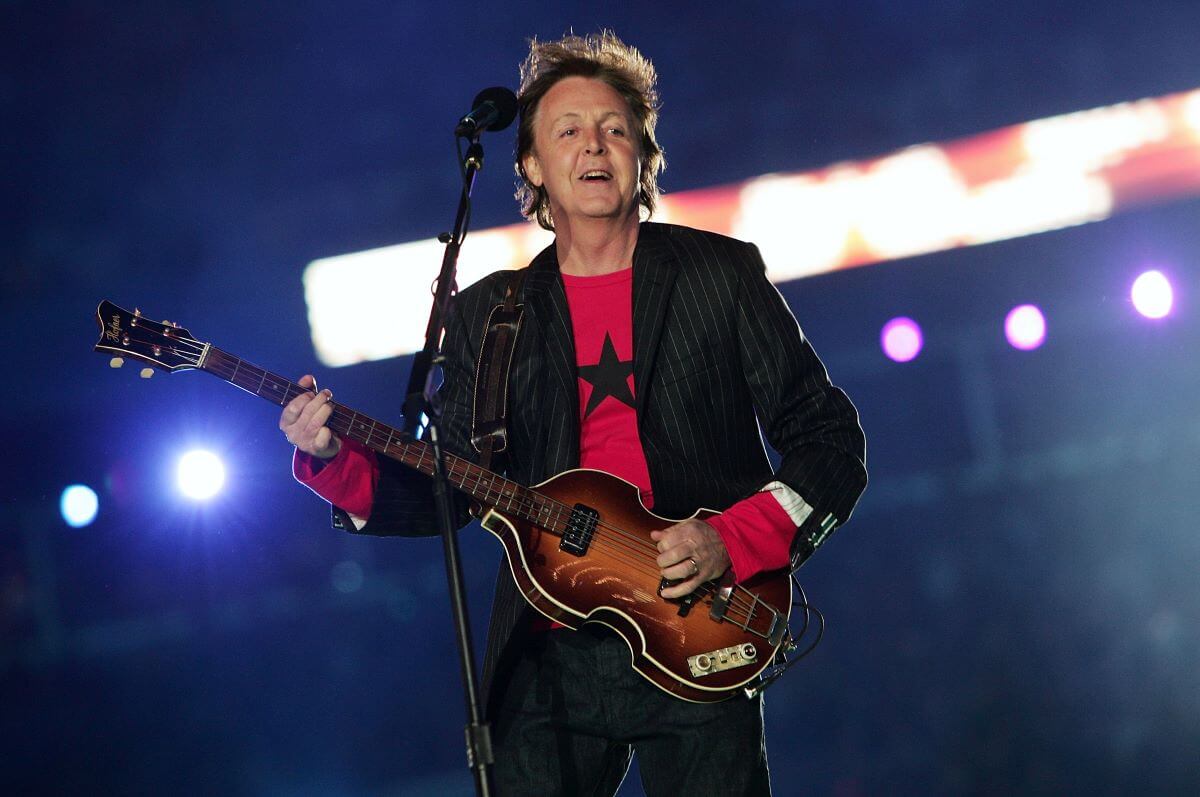 Paul McCartney wears a red shirt and black blazer. He plays guitar.