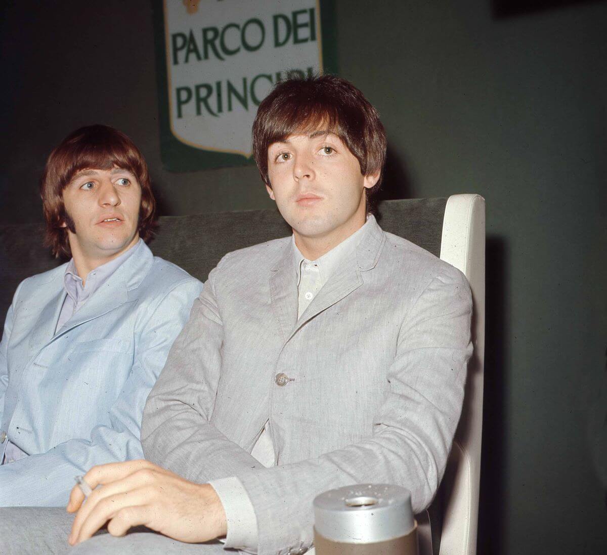 Ringo Starr and Paul McCartney sit together on the same side of a table. They wear gray suits.