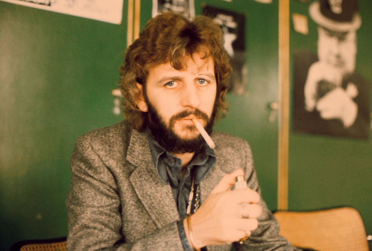 Ringo Starr sits in front of a green wall and holds a cigarette in his mouth. There is a lighter in his hand.