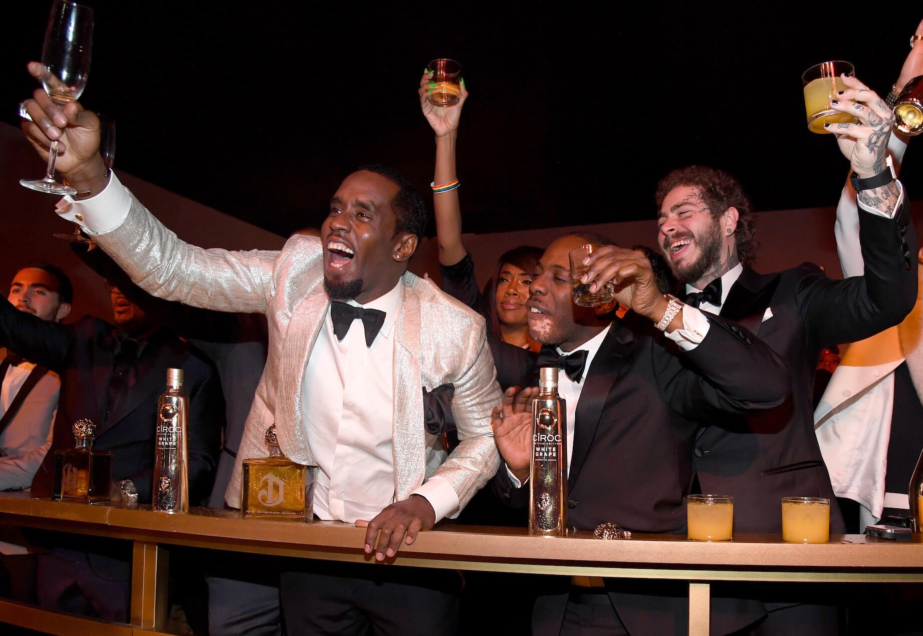 Sean 'P. Diddy' Combs wearing a white suit and holding up a drink for his 50th birthday. Post Malone and other celebrities raise their drinks in the background.