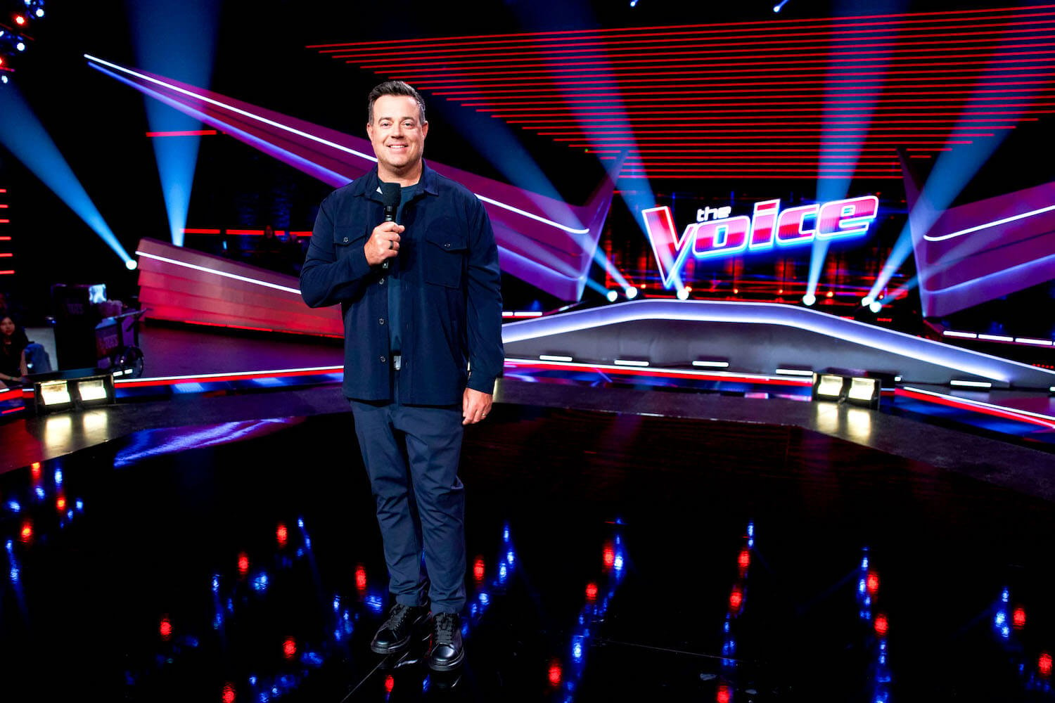 Carson Daly holding a microphone with 'The Voice' logo in the background