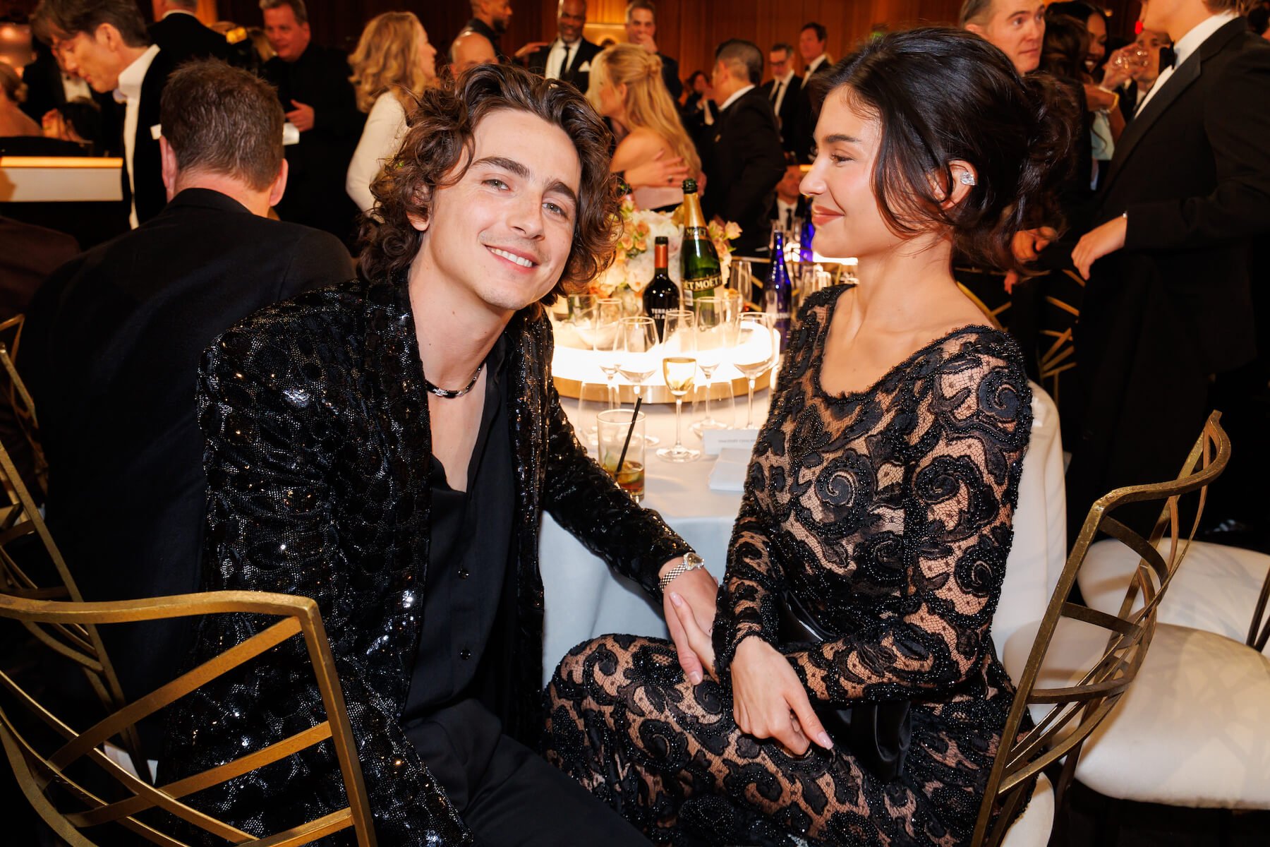 Timothée Chalamet sitting at a table at the Golden Globes and smiling at the camera while Kylie Jenner looks at him and smiles